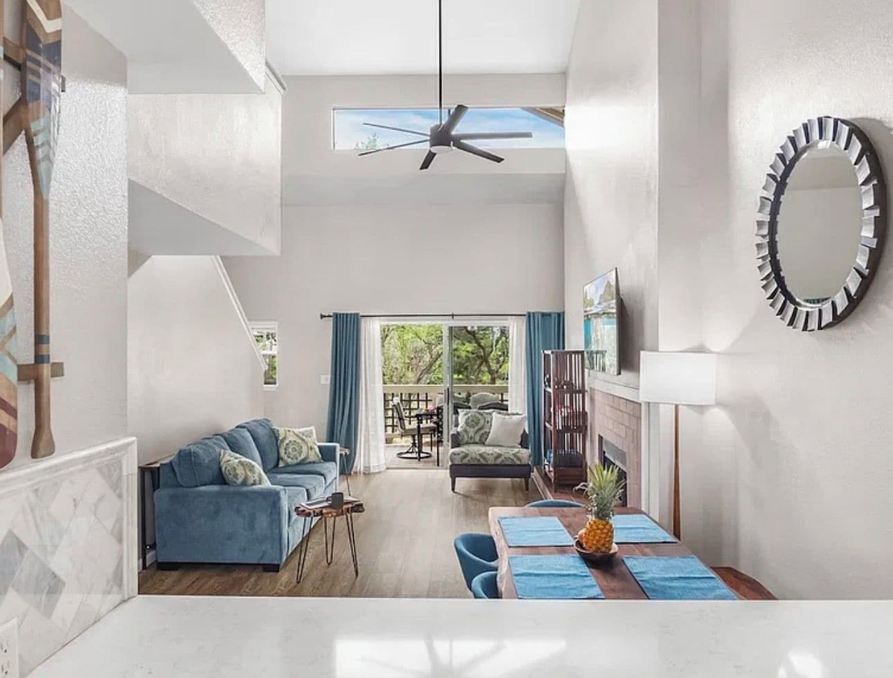 Ein geräumiger Wohnbereich mit Deckenventilator, einem Esstisch mit blauen Platzsets und Blick durch eine Glastür auf eine Außenpatio.