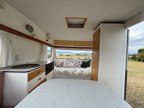 Een compact RV-interieur met een kitchenette, een bed met kussens, en een raam met uitzicht op de natuur.