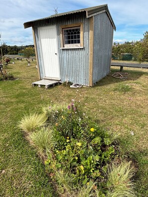A small, single-story metal shed with a window and a door, situated on a grassy area with a garden bed in the foreground. - The Stoney Shore - boutique lakeside camp. Breathtaking lake mountain views (Te Anau)
