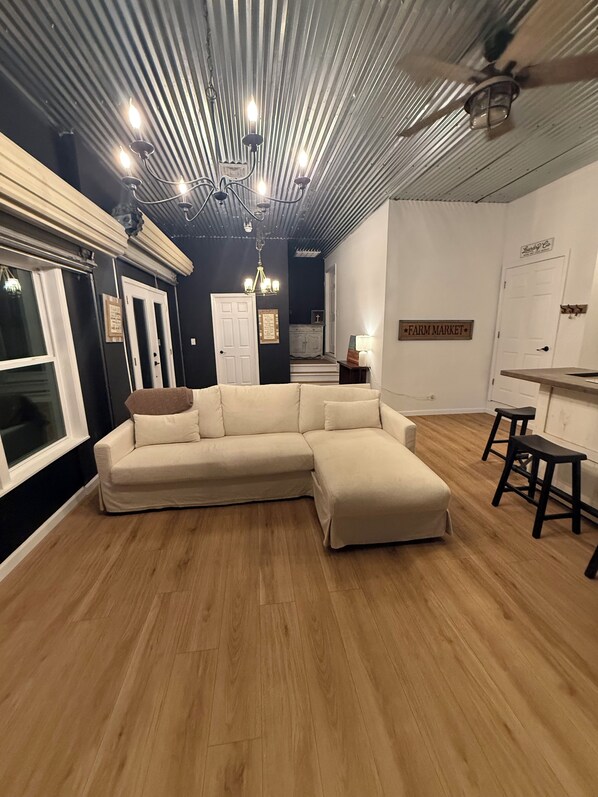 Una habitación con sofá modular, suelo de madera, ventilador de techo y una zona de cocina con taburetes.