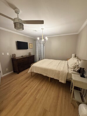 Un dormitorio con cama, cómoda, televisor, candelabro, ventilador de techo y un escritorio con lámpara.