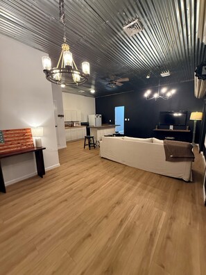 A modern living room with a white sofa, a wooden floor, a chandelier, and a kitchen area in the background.