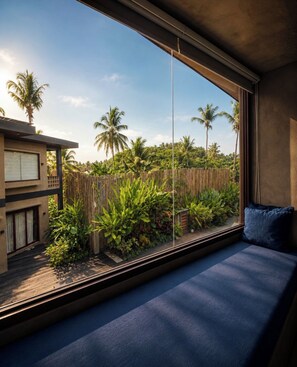 Ein Zimmer mit großem Fenster und Blick auf einen tropischen Garten mit Palmen und einem Holzzaun.