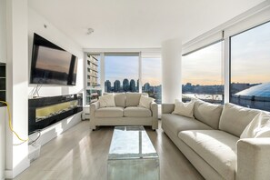 A modern living room with a large TV, two sofas, a coffee table, and a city view through large windows. - Bright 2BR Condo w/ Pool Across from Stadium (Vancouver)