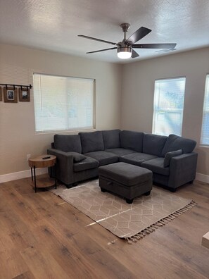 A living room with a sectional sofa, a small side table, a ceiling fan, and a patterned rug. - The Valley Haus (Sanger)