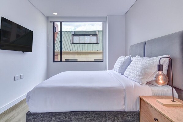A modern bedroom with a bed, a nightstand, a lamp, and a TV mounted on the wall. - New Listing (Bogotá)