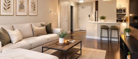 A modern living room with a sofa, coffee table, and a kitchen area with a microwave and bar stools.