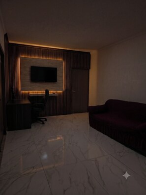 A room with a flat-screen TV mounted on a wall, a dark-colored sofa, a small desk with a chair, and a tiled floor. - New Listing (Guadalupe)