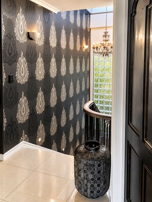 A hallway with patterned wallpaper, a chandelier, and a decorative urn. - Palms on Tindalls Beach side retreat (Whangaparāoa)
