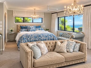 A bedroom with a large bed, a chaise lounge, a chandelier, and a view of the outdoors. - Palms on Tindalls Beach side retreat (Whangaparāoa)
