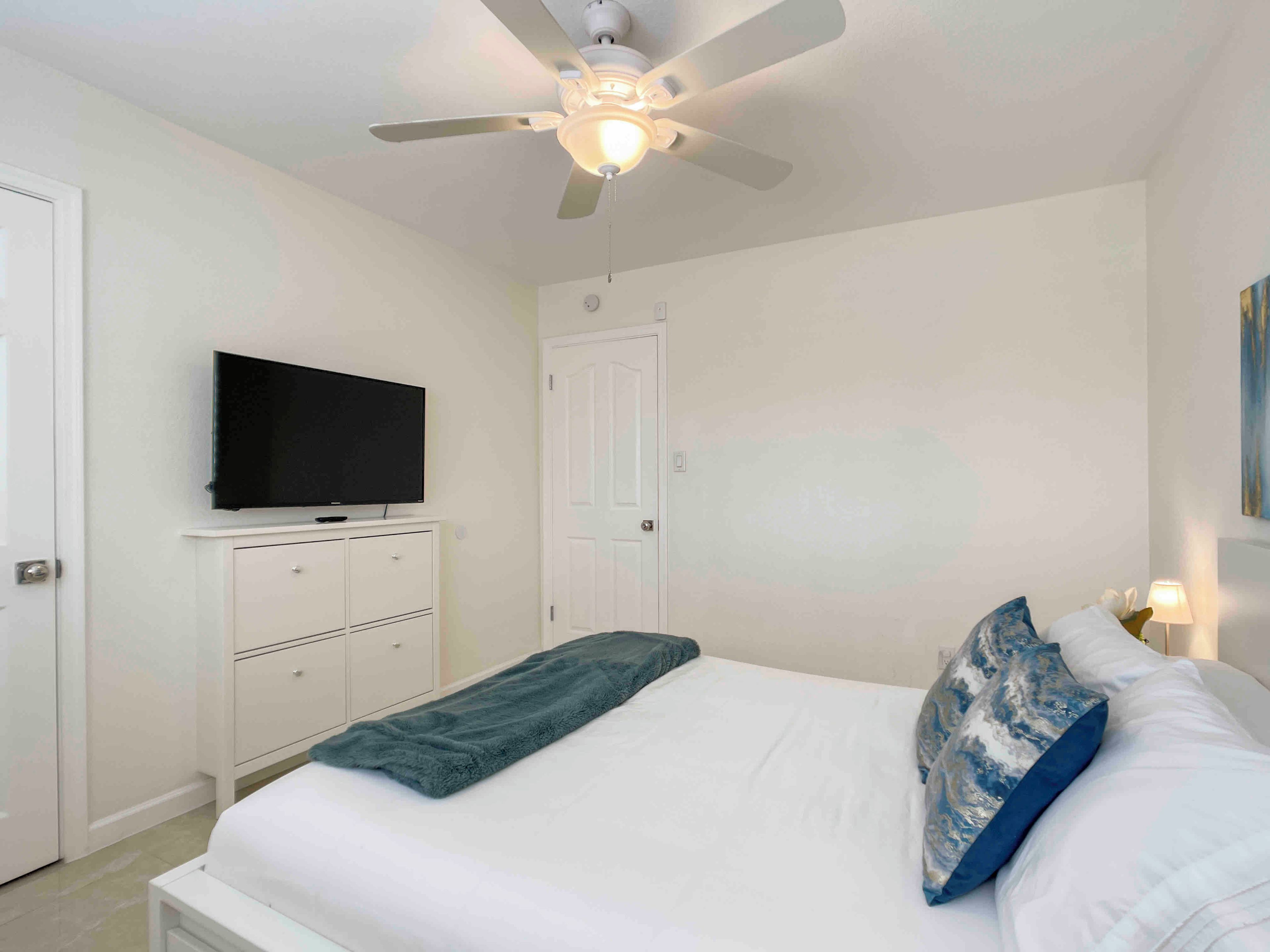 Un dormitorio con una cama, un ventilador de techo, un televisor sobre un mueble y una puerta.