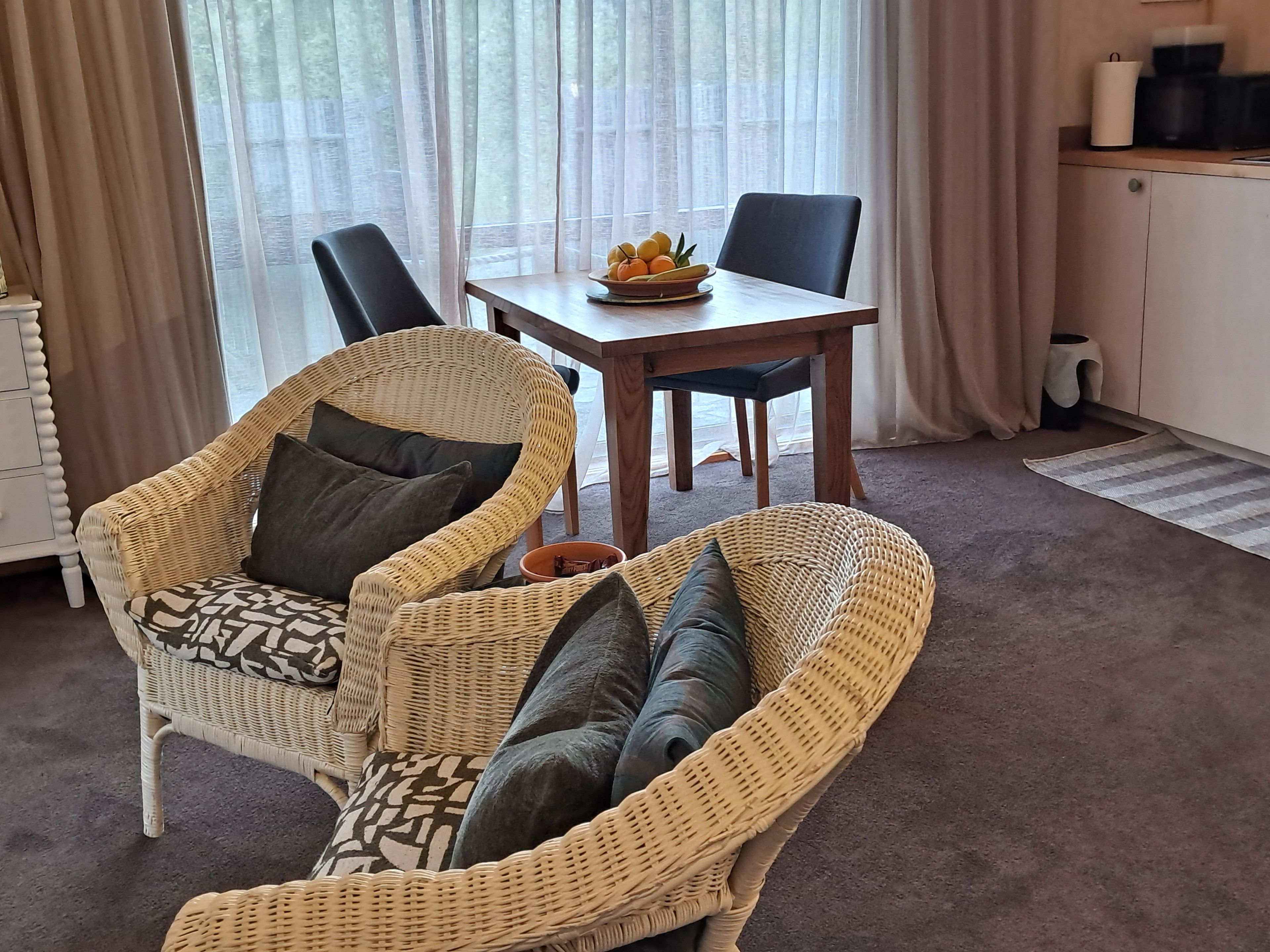 A room with a wicker sofa, a dining table with a fruit bowl, and a kitchen area with a coffee maker.