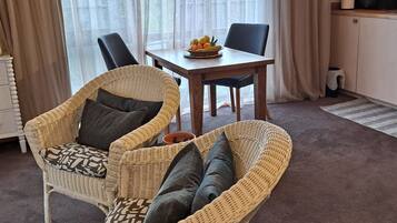 A room with a wicker sofa, a dining table with a fruit bowl, and a kitchen area with a coffee maker.