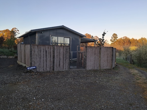 A small, single-story building with a wooden fence, a clear sky, and trees in the background.