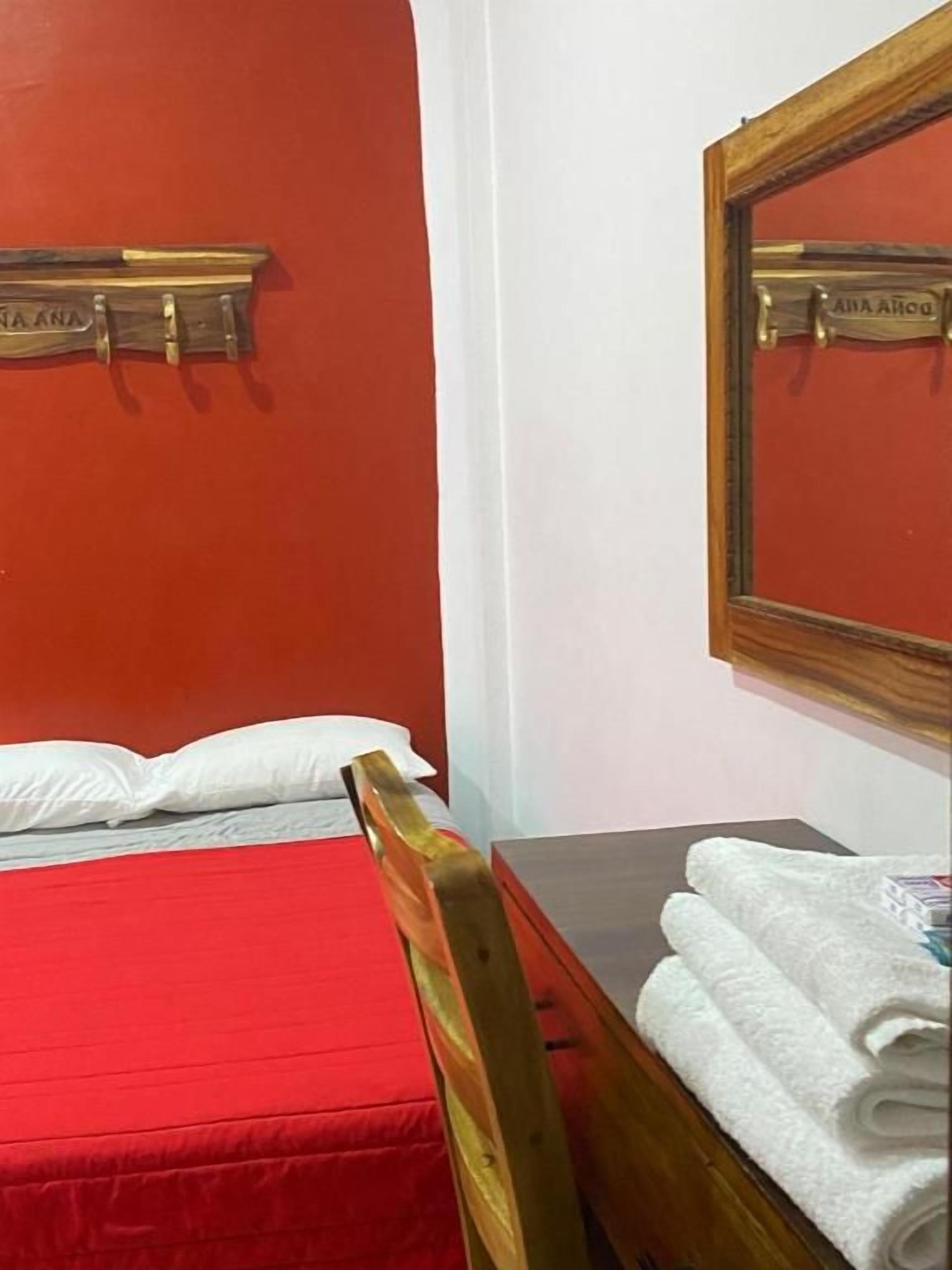 Una habitación con una cama, una silla de madera, una mesa con toallas y un espejo con una placa de identificación.