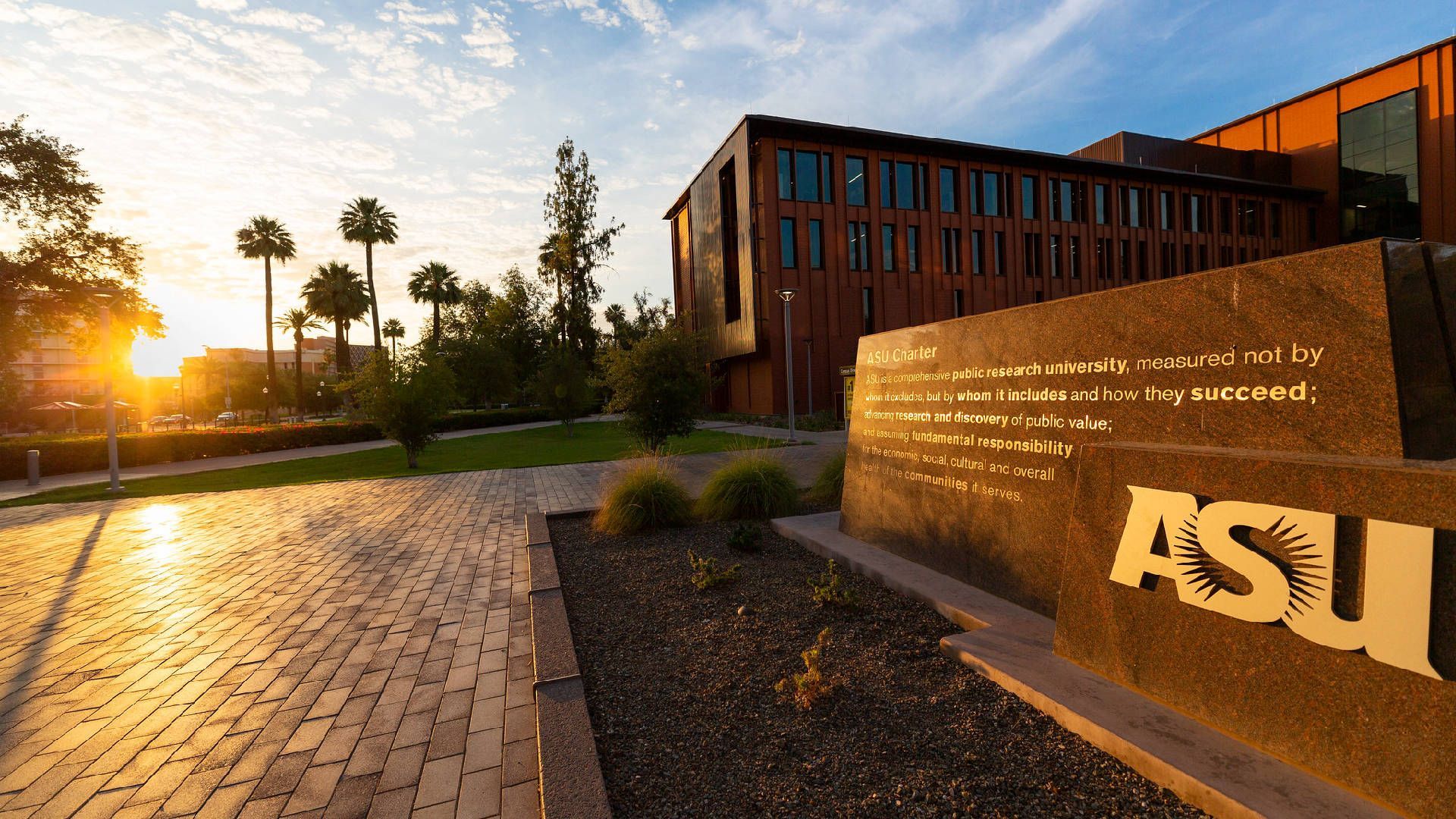 A university campus with a modern building, a paved walkway, and a sign with the university's name.