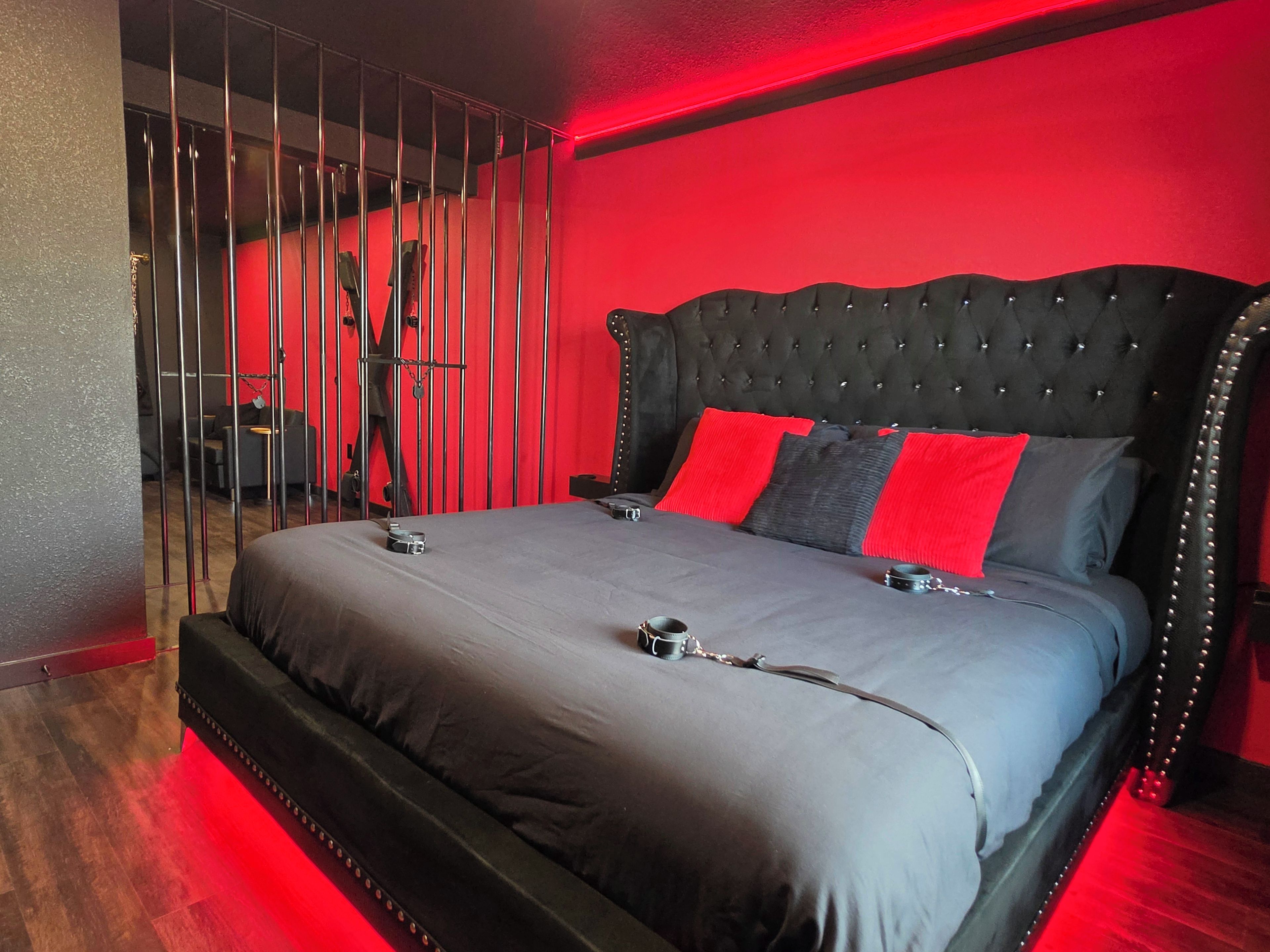 A bed with a black headboard, grey bedding, and red pillows in a room with red walls and a red-lit floor.