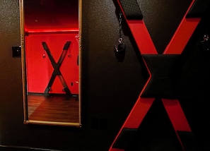 A room with a mirror reflecting a red-lit space, a black and red X-shaped decoration, and a black strap hanging on the wall. - NEW! Red Room. Spicy Couples Escape.  Clean!  King Bed. Near WinStar.  Private. (Thackerville)