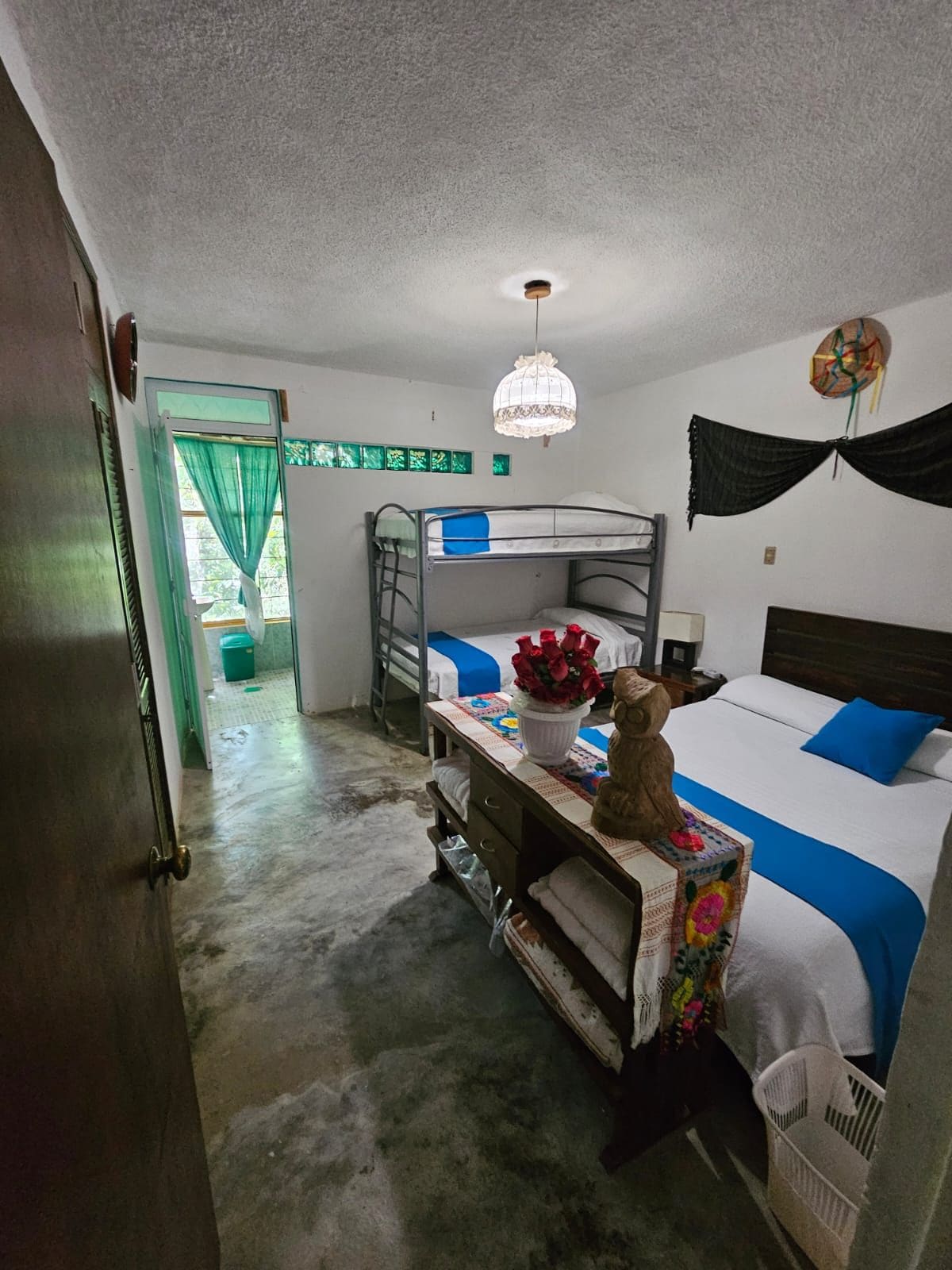 A room with bunk beds, a desk with a chair, a hanging light fixture, and a door leading to another room.