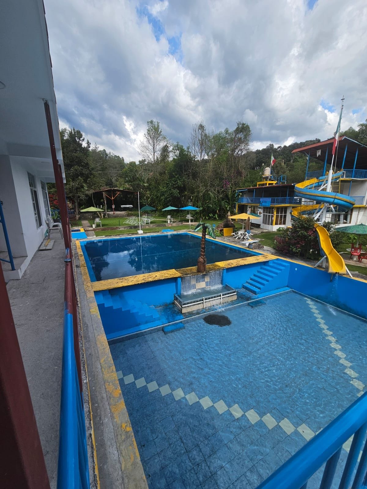 A swimming pool with a slide, surrounded by a building with a red roof and a view of trees and hills in the background.