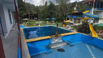 A swimming pool with a slide, surrounded by a building with a red roof and a view of trees and hills in the background.