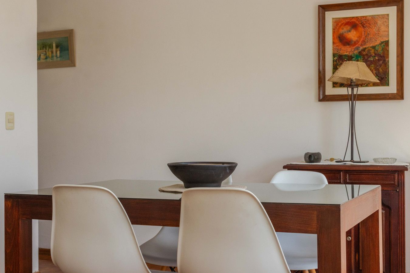 A dining area with a wooden table, two white chairs, a black bowl, a lamp, and a framed painting on the wall.