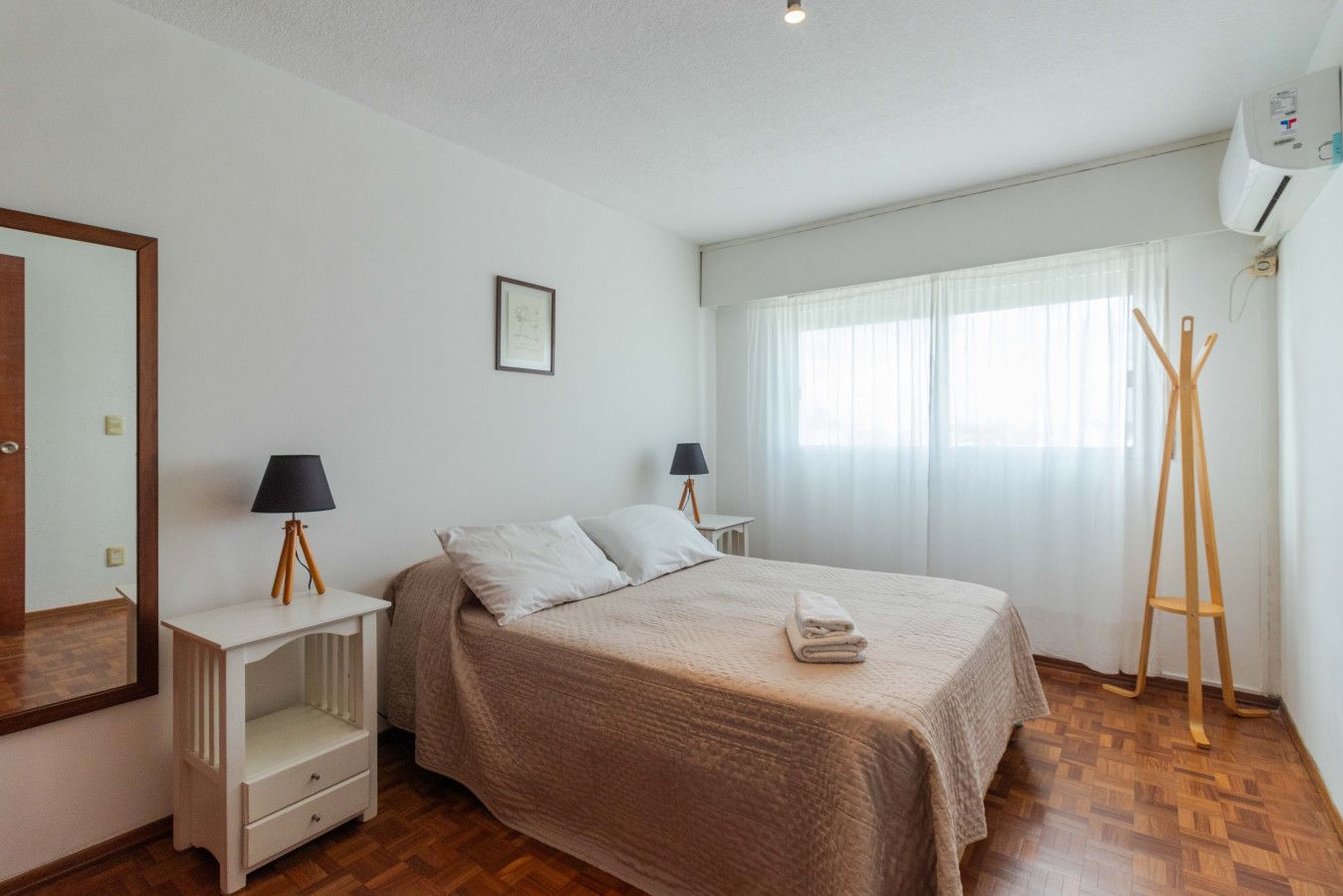 Ein Schlafzimmer mit einem Bett, zwei Nachttischen, einem Spiegel, einem Fenster mit Vorhängen und einer Klimaanlage.