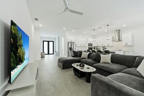 A modern living room with a flat-screen TV, grey sofas, a coffee table, and a kitchen in the background. - 4BR All-Suite Home, 5.5 Baths, Heated Pool & Spa (Bonita Springs)