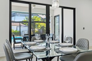 A dining area with a glass table set for four, surrounded by chairs, with a view of a swimming pool and greenery outside. - 4BR All-Suite Home, 5.5 Baths, Heated Pool & Spa (Bonita Springs)
