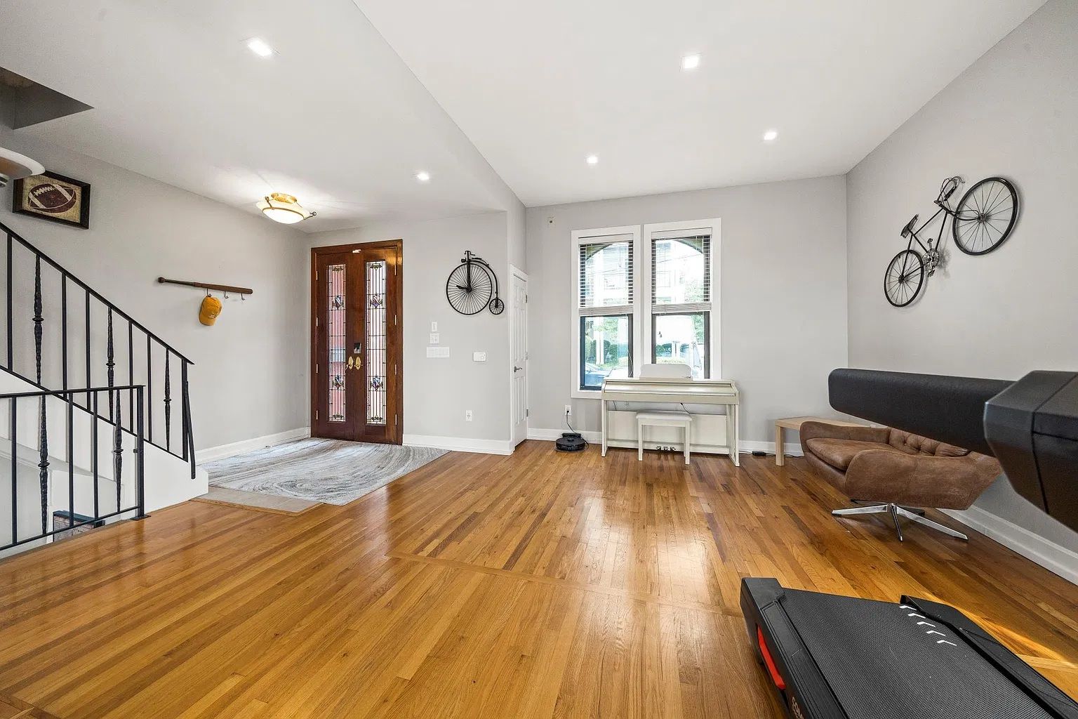 A spacious living room with hardwood flooring, a piano, a brown leather couch, and a treadmill.