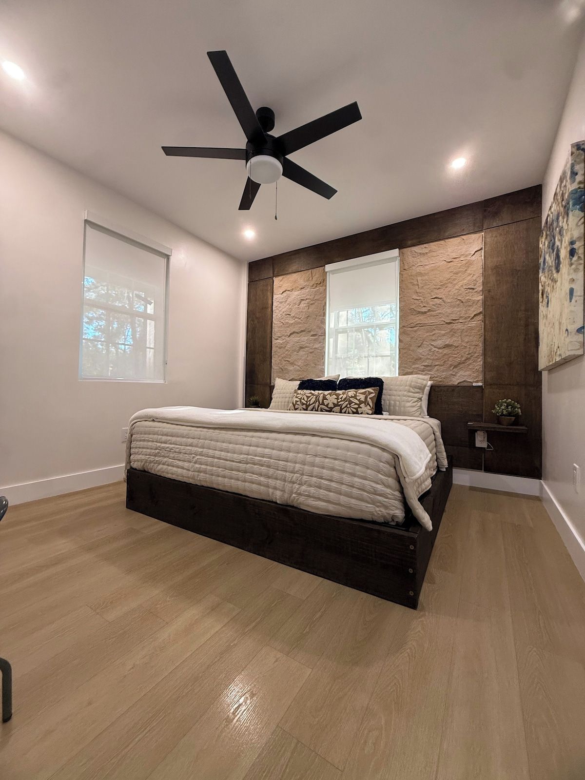 A bedroom with a bed, a ceiling fan, a window, and a wall-mounted shelf.