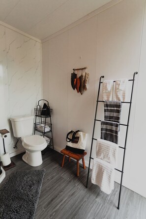 A bathroom with a toilet, a towel rack, a shelf with bags, and a small stool.