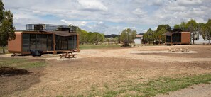 Two modern, brown, glass-enclosed cabins with picnic tables, set in a dry, grassy area with trees and a distant hill. - Glass Cave King unit at WYLD ARBOLES near Navajo lake. Pet Friendly! (Ignacio)