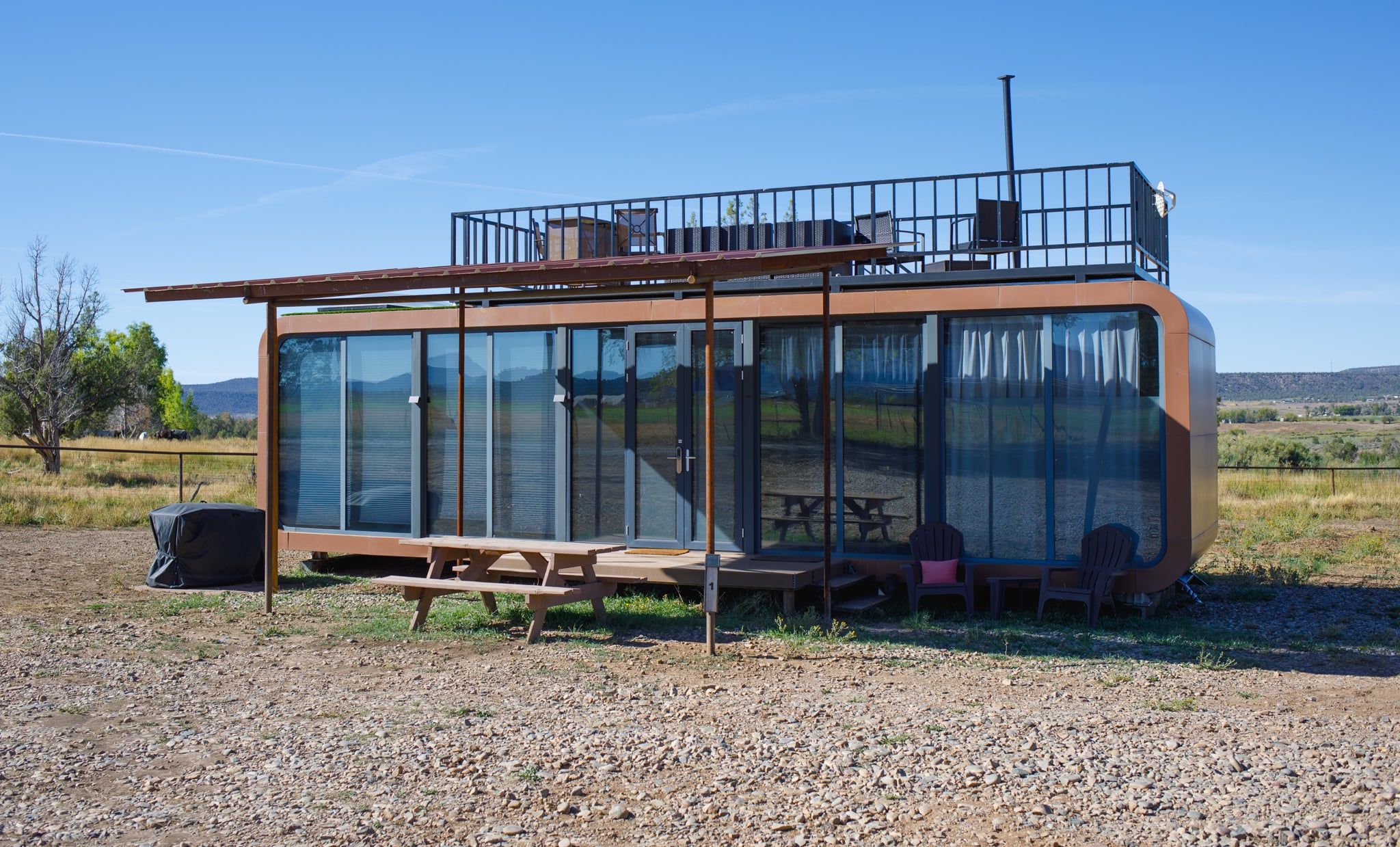 A modern, glass-enclosed structure with a balcony, outdoor seating, and a gravel ground.