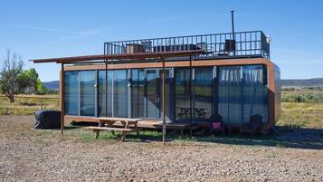 A modern, glass-enclosed structure with a balcony, outdoor seating, and a gravel ground.