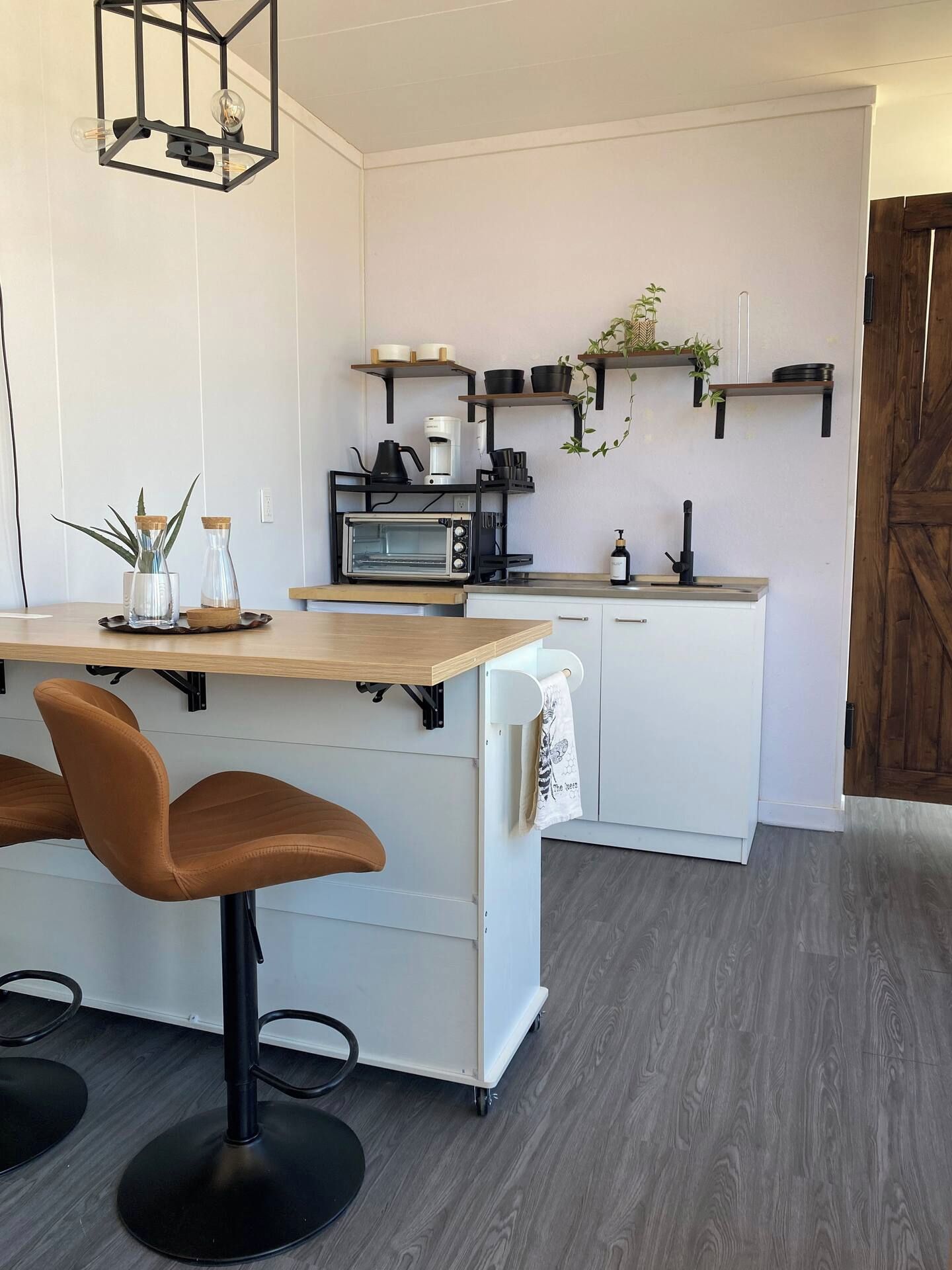 A modern kitchen with a central island, bar stools, a microwave, a coffee maker, and hanging plants.