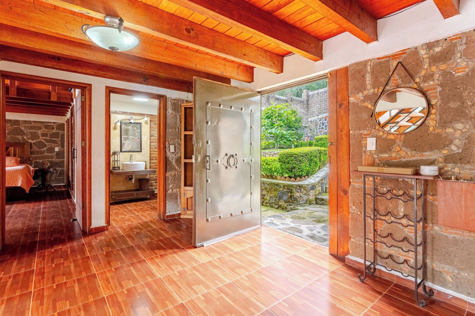 A spacious room with a wooden ceiling, a large open door leading to a bathroom, and a stone wall with a circular mirror.
