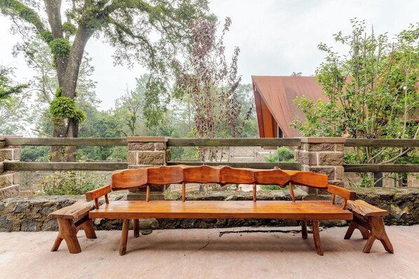 A wooden bench with a stone wall backdrop, surrounded by greenery and a building with a red roof. - Forest cabin for groups · hot tub and fire pits (Tres Marias)