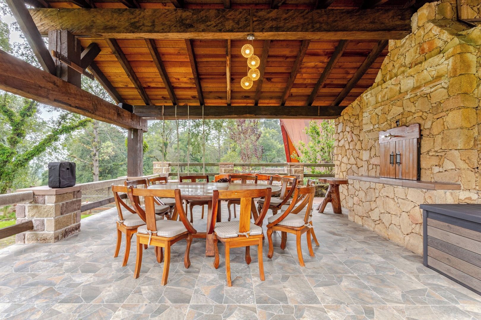 An outdoor patio with a stone floor, wooden tables and chairs, a stone wall, and a wooden cabinet.