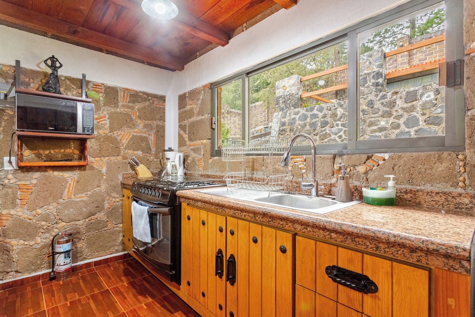 A kitchen with a stone wall, wooden cabinets, a stainless steel sink, and a window with a view of greenery.