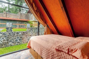A bedroom with a large window, a stone wall outside, and a bed with a pink bedspread. - Forest cabin for groups · hot tub and fire pits (Tres Marias)
