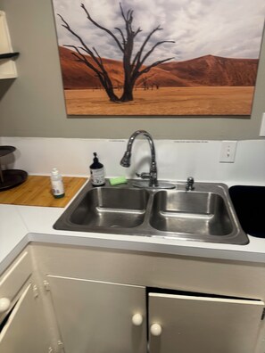 A kitchen with a double stainless steel sink, a wooden cutting board, and a bottle of hand sanitizer. - Cozy 1-bedroom apartment in delightful Houston with AC in the Midtown Area (Houston)