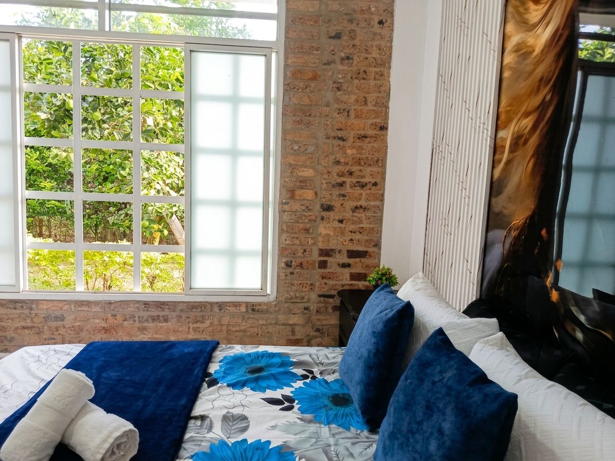 A room with a brick wall, a large window with a view of greenery, a bed with blue and white bedding, and a white headboard.