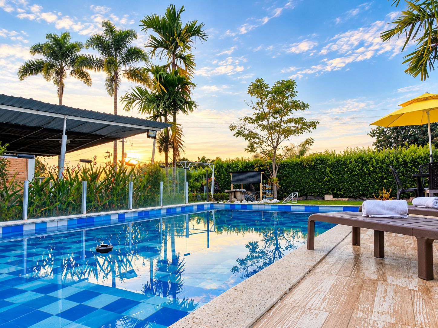 A swimming pool with a tiled deck, surrounded by palm trees and a covered seating area.