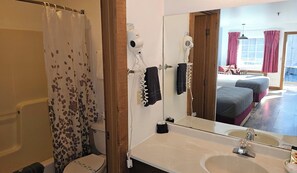 Un baño con ducha, inodoro, secador de pelo, toallero y espejo que refleja un dormitorio con cama y cortinas.