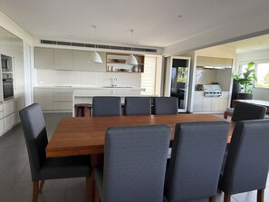 A modern kitchen with a central island, dining area with a wooden table and chairs, and a view of the outdoors. - Ocean views everywhere, walking distance to town. (Lennox Head)