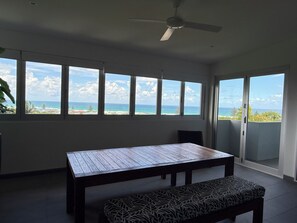 A room with a large table and bench, a ceiling fan, and a view of the ocean through large windows. - Ocean views everywhere, walking distance to town. (Lennox Head)