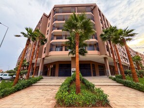 Un edificio de varios pisos con una entrada central, rodeado de palmeras y un sendero pavimentado.