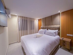 Hotellihuone, jossa on sänky, yöpöytä, seinälle asennettu televisio ja ikkuna, jossa on verhot.