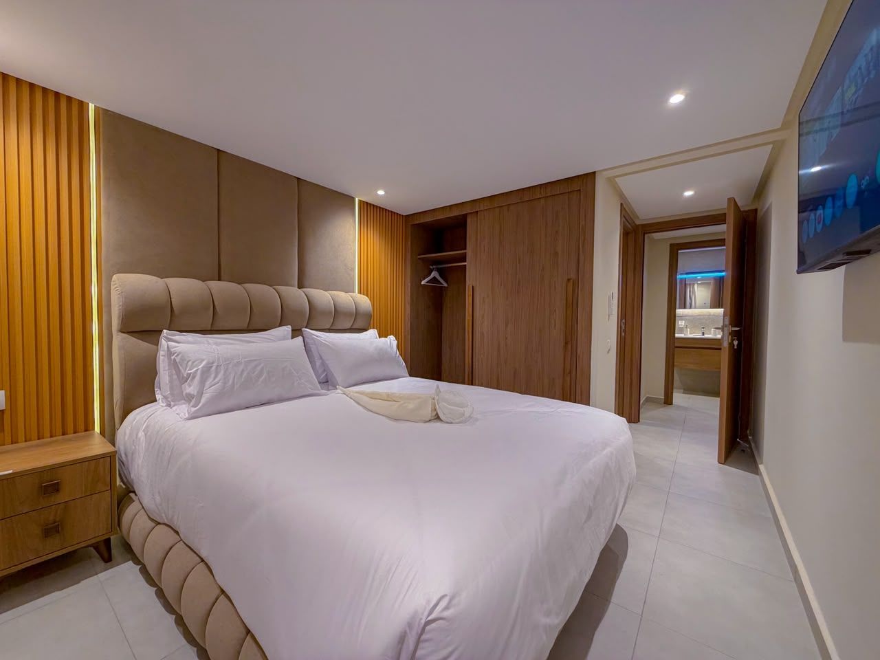 A modern bedroom with a large bed, wooden paneling, a bedside table, and a TV mounted on the wall.
